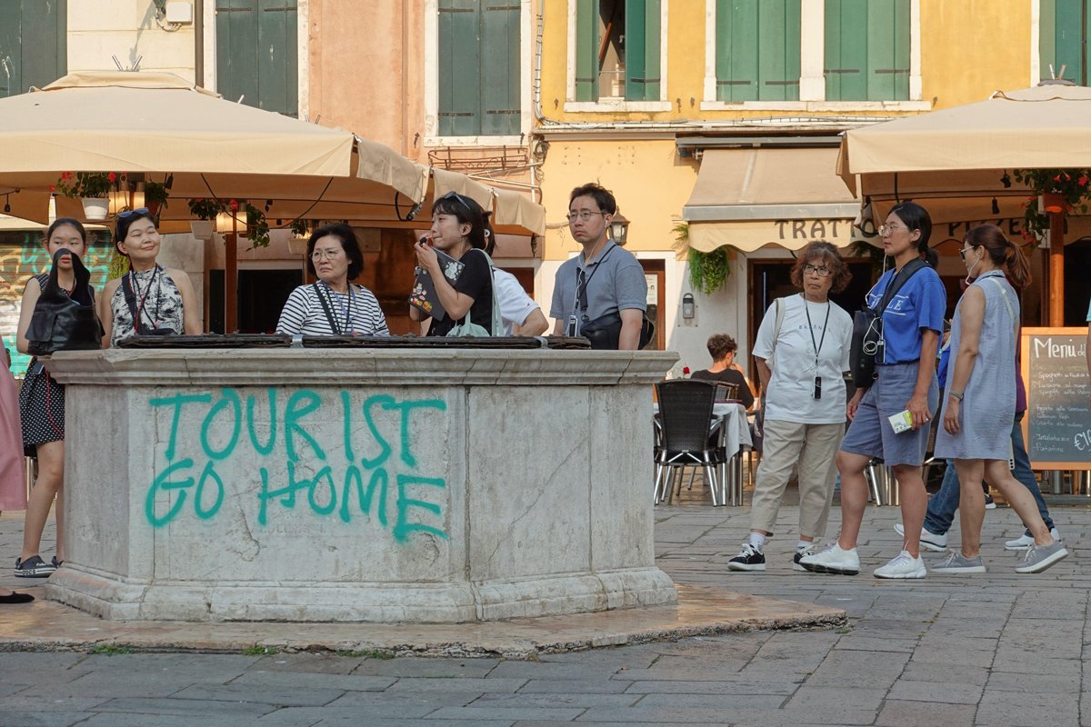 Туристы на фоне графити «Tourist Go Home» в Венеции, июнь 2024 года. Фото: Viennaslide / Alamy / Vida Press